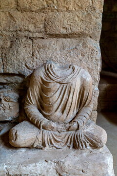 View of a weathered, headless Buddha statue sits serenely against a rough stone wall, exuding ancient peace, tactile textures, and soft light, Taxila, Khyber Pakhtunkhwa, Pakistan.