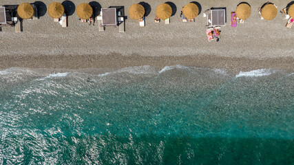 Top-down aerial shot capturing the meeting point of land and sea. Aerial perpendicular view of a single row of golden straw parasols and beach cabanas lines the pebble shore. It is a sunny summer day. © Stefano Tammaro
