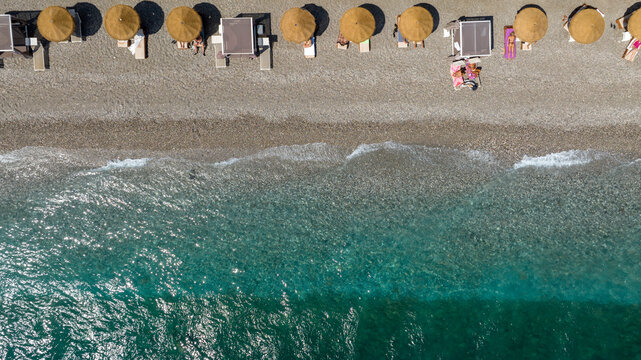Top-down aerial shot capturing the meeting point of land and sea. Aerial perpendicular view of a single row of golden straw parasols and beach cabanas lines the pebble shore. It is a sunny summer day.