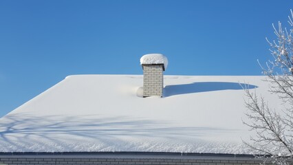 Color photo of a house completely covered in snow, with icicles hanging from the roof and chimney, set against a backdrop of snowy trees and a clear blue sky.