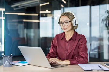 Businesswoman in a modern office setup wearing headphones, intensely focusing on her laptop,...