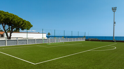 Panorama of an artificial turf football pitch overlooking the sea. The vibrant green field is framed by a clear blue sky and a deep Mediterranean horizon, featuring a modern fence and floodlights.