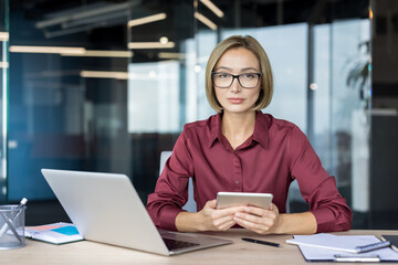 Young businesswoman in glasses sits at a modern desk with laptop and tablet, looking confidently at...