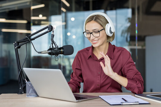 Young woman smiling, wearing headphones and glasses, waving while recording a podcast or participating in a live online broadcast from her modern home studio setup with a microphone and laptop