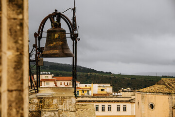 Historic Bell Overlooking Ragusa Cityscape In Sicily: Weathered Bronze Bell Framed Against Stone Buildings, Tiled Rooftops, And Overcast Sky