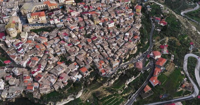 Aerial view of buildings with red roofs, nestled closely together, contrasting with the winding roads and green vegetation, Agira, Sicilia, Italy.
