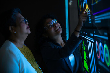 Two diverse professional women analyzing complex data charts on large futuristic digital screen. Senior executive and young analyst collaborating in dark high-tech office with blue neon interface.