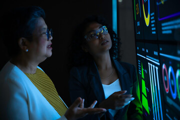 Two diverse professional women analyzing complex data charts on large futuristic digital screen. Senior executive and young analyst collaborating in dark high-tech office with blue neon interface.