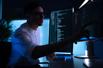 Focused software developer working late at night in dark office. Professional programmer coding on vertical monitor with blue neon light. Cybersecurity expert analyzing data alone.