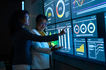 Two diverse professional women analyzing complex data charts on large futuristic digital screen. Senior executive and young analyst collaborating in dark high-tech office with blue neon interface.
