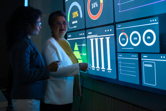 Two diverse professional women analyzing complex data charts on large futuristic digital screen. Senior executive and young analyst collaborating in dark high-tech office with blue neon interface. - Powered by Adobe