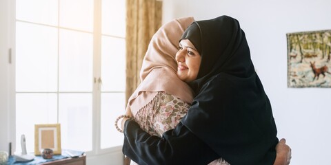 Muslim women in hijabs embrace warmly in a sunlit room. Friendship and connection are highlighted as they share a heartfelt hug, symbolizing unity and support. Two Muslim women greeting with hug.