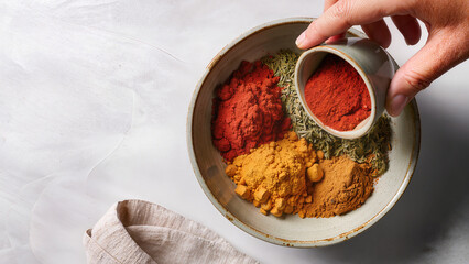 Human hand adding red spice to mix of colorful ground spices in ceramic bowl on light background. Concept of culinary creativity, healthy cooking, natural ingredients, and food styling