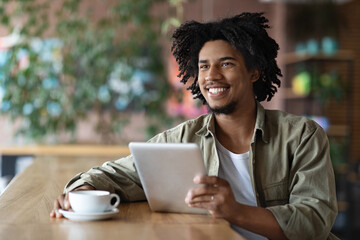 Smiling millennial black curly male blogger with tablet sitting at table, drinking coffee and look at free space in cafe interior. Modern technology, work remotely, chat in social networks and app