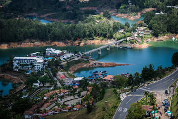 View of the vibrant reservoir reflecting the sky with lush green hills, a bridge connecting land, and buildings dotting the landscape, Guatape, Antioquia, Colombia.