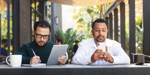 Two men working outdoors. One man writes notes, the other checks his phone. Casual work setting with coffee. Outdoor work, notes, phone, coffee. Two men working in an outdoor cafe.