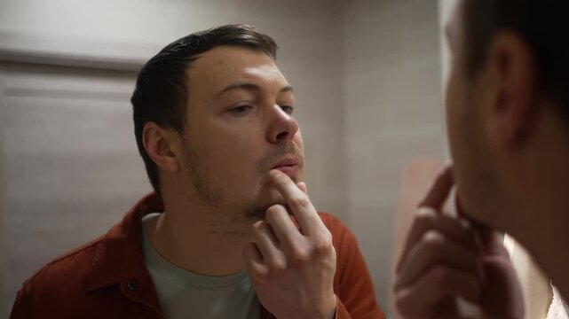 Young man standing in a bathroom and looking in the mirror, carefully applying medicinal ointment with his finger to a cold sore on his lip, a manifestation of the herpes simplex virus