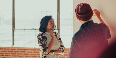 Two diverse people in a casual setting, one writing on a board, the other observing. Bright office with open space and large windows with a relaxed atmosphere.