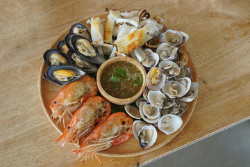 top view hot grilled seafood, three large shrimps, cut squid, cockles, scallops and seafood dipping sauce in a wooden bowl on a wooden tray, on a wooden table background