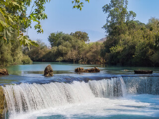 Manavgat Waterfall, Turkey. Stunning wide view of the famous turquoise water cascading over a low rocky ledge, surrounded by lush green trees under a bright sunny sky.