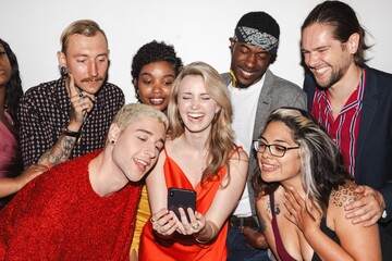 Diverse group of friends laughing, gathered around a smartphone. Mixed ethnicities, men and women, enjoying a fun moment together, sharing smiles and joy. Diverse friends taking selfie at party.