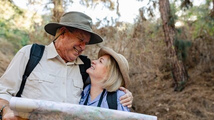 Elderly couple hiking with a map. Smiling elderly man and woman hiking together. Elderly hikers enjoying nature. Elderly couple exploring outdoors. Elderly man holding map and embrace his wife
