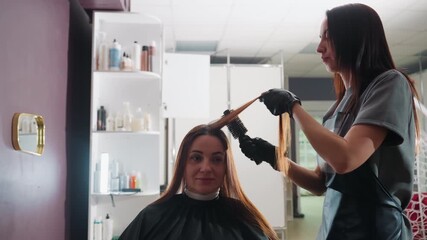 colorist applies dye to client hair with brush and gloves, careful sectioning and smoothing, bowls and developer on nearby shelves, professional cape and focused technique under bright salon lights
