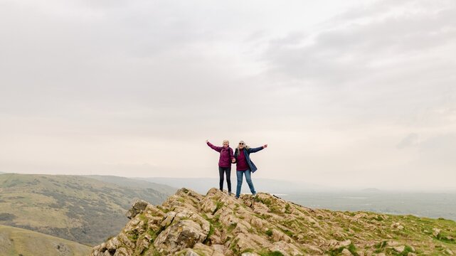 Two senior people stand on a rocky hilltop with arms outstretched, enjoying the view. Overcast sky, rocky terrain, and nature landscape. Two seniors enjoying the mountain view. Seniors travelling.
