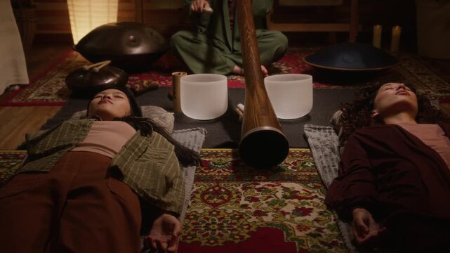 Medium shot of two diverse young women lying comfortably on carpet in studio, meditating and enjoying relaxing ambiance, while female practitioner is playing didgeridoo and singing bowls