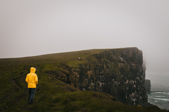 View of a solitary figure in a bright yellow raincoat walks along a grassy path towards the edge of a dramatic cliff face shrouded in mist, Latrabjarg, Westfjords, Iceland.