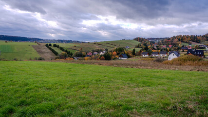 A tranquil wide-angle view of a charming rural village nestled among rolling green hills and vibrant autumn foliage under an overcast sky.