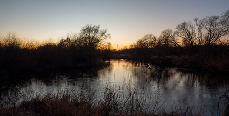 A serene river meanders through a tranquil winter landscape at twilight, featuring bare trees and reeds reflected in calm, still waters under a peaceful sky.