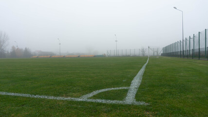 Empty soccer pitch shrouded in thick, atmospheric fog, featuring crisp white boundary lines and a surrounding fence on a quiet, isolated morning.