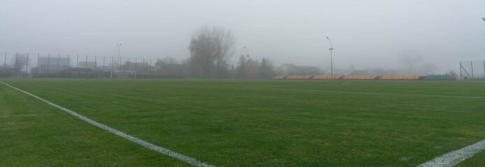 An empty green sports field blanketed in thick, gray fog under a muted overcast sky, conveying a...