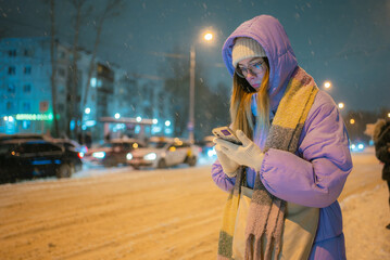 Young woman checking phone for bus schedule while standing at a snowy winter street bus stop during...