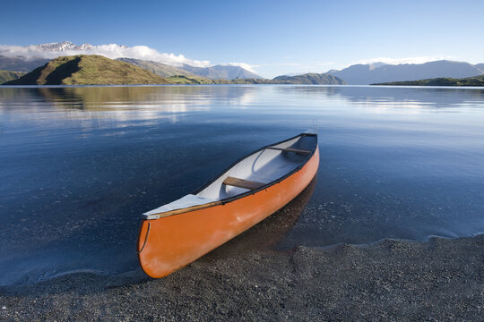 View of a vibrant orange canoe resting on the dark, grainy shore of a tranquil lake mirroring snow-capped mountains under a clear blue sky, Wanaka, Otago Region, New Zealand.