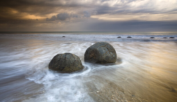 View of spherical Moeraki Boulders stand defiant against the surging tide under a dramatic sky, a timeless dance of rock and water, Hampden, Otago Region, New Zealand.
