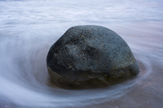 View of a solitary, weathered boulder embraced by the swirling, ethereal dance of ocean waves on a sandy shore, Hampden, Otago Region, New Zealand.