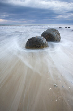 View of spherical Moeraki Boulders stand defiant against the relentless surge of milky white waves under a brooding sky, Hampden, Otago Region, New Zealand.