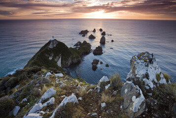 View of rugged cliffs meeting the vast ocean under a vibrant sunset sky, with a distant lighthouse standing sentinel, Kaka Point, Otago Region, New Zealand.