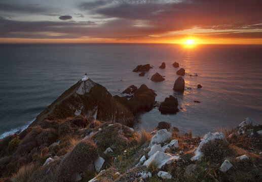 View of golden light bathing the Nugget Point Lighthouse atop the rugged headland, casting long shadows on the turquoise sea, Kaka Point, Otago Region, New Zealand.