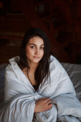 A woman with black hair wakes up in the morning in a white blanket and catches the rays of the sun. The portrait is creative, with brown eyes. High quality photo