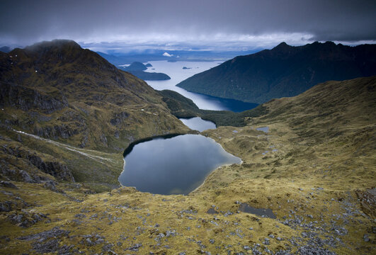 View of dark waters mirroring the moody sky, rugged mountains cascading down to meet the serene lake, a stark contrast of textures and tones, Christchurch, West Coast Region, New Zealand.