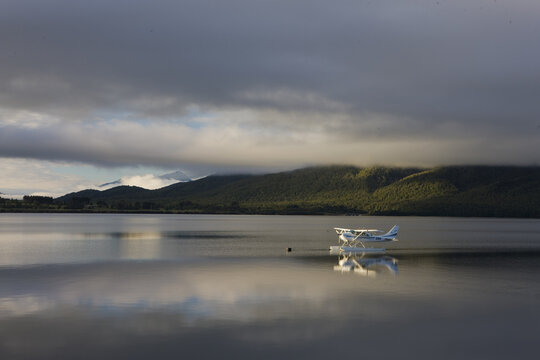 View of a seaplane rests on the tranquil waters, mirroring the cloudy sky and distant green hills, creating a serene contrast, Christchurch, West Coast Region, New Zealand.