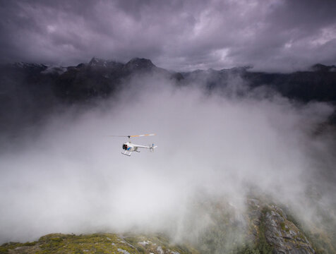 View of a helicopter gracefully soars amidst a sea of ethereal clouds, dwarfed by the majestic, dark mountains under a stormy sky, Christchurch, West Coast Region, New Zealand.