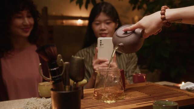 Medium shot of multiethnic girlfriends watching traditional gongfu ceremony at tea house, while unrecognizable master pouring hot infusion from teapot into pitcher, and filming on smartphone