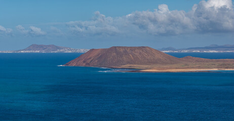 Aerial view shows a rust colored caldera and lava fields by Fuerteventura, pale sandy beaches, thin surf, deep Atlantic waters, and Lanzarote settlements under clear midday light. © Aerial Film Studio