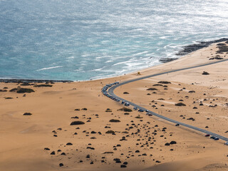 Obraz premium Aerial view of Fuerteventura, Canary Islands, with coastal road, parked cars, dunes, volcanic rock, and turquoise Atlantic in late afternoon near Morro Jable and Jandia.