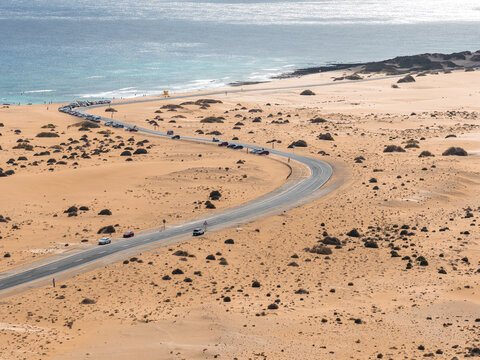 Aerial view shows a curving road through golden dunes by the Atlantic in Fuerteventura, Canary Islands, cars heading to Playa del Bajo de la Burra under soft daylight.