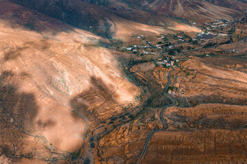 Aerial arid hills and terraced valleys on Fuerteventura, Canary Islands, with a sinuous road, whitewashed houses, patchy vegetation, and cloud shadows in warm daylight © Aerial Film Studio
